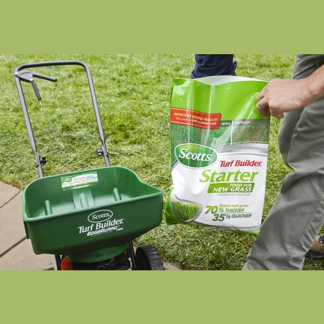 Person holding a bag of lawn starter fertilizer next to a green Scotts spreader on grass in plant care collections