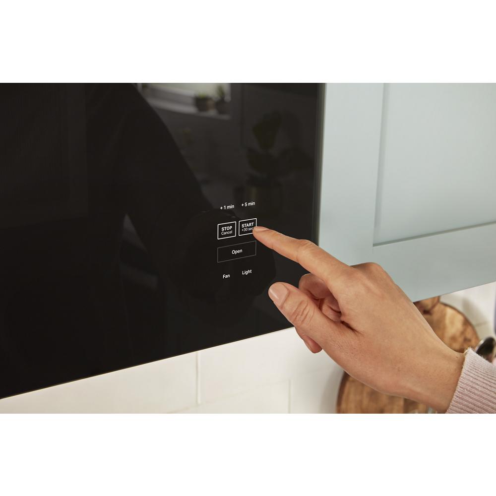 Hand pressing start button on sleek black glass control panel of built-in microwave in modern kitchen setting