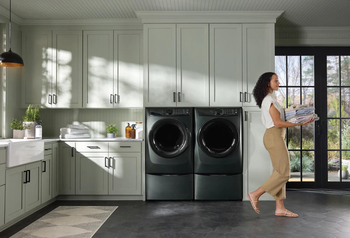 Modern laundry room with dark gray front load washers and woman carrying folded linens near large window