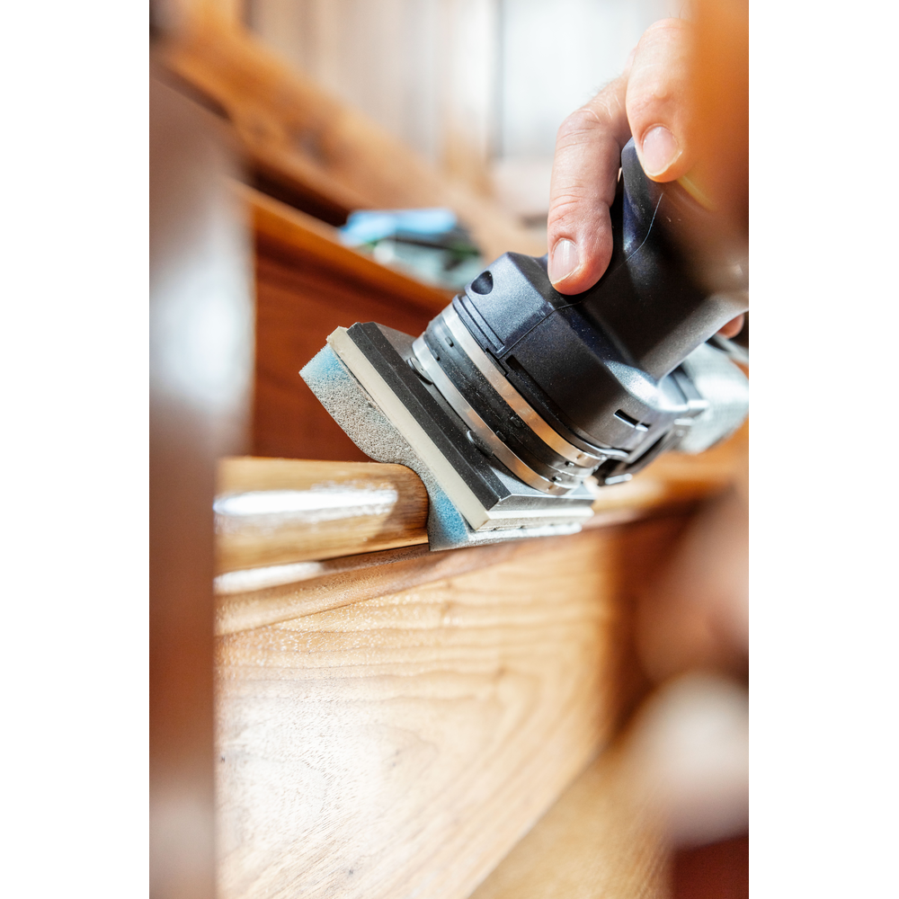 Person operating a handheld power tool with foam-backed abrasive smoothing wooden stair detail in home improvement setting