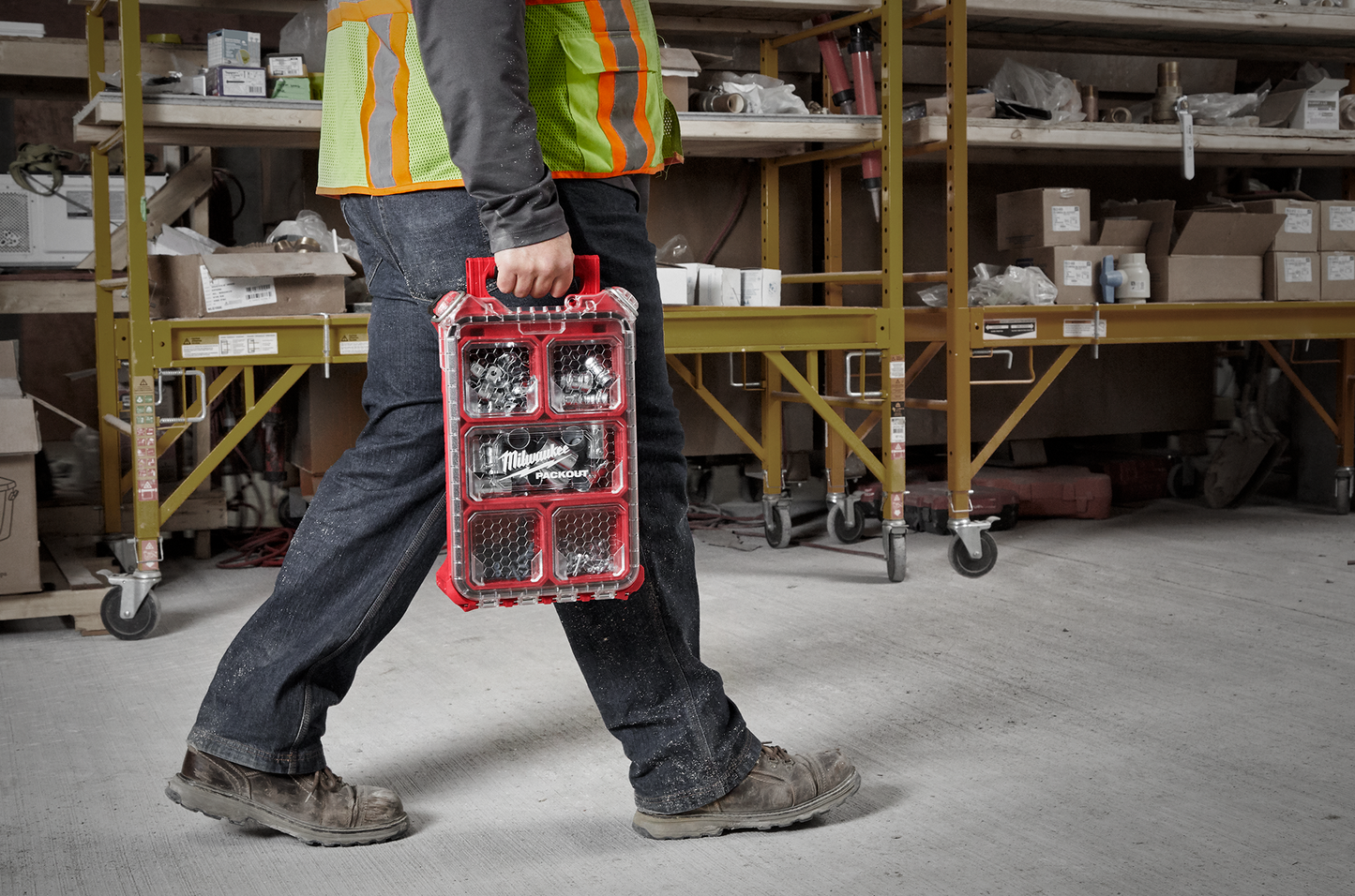 Person wearing safety vest carrying red Milwaukee low-profile compact organizer with clear lid and multiple compartments for tool storage