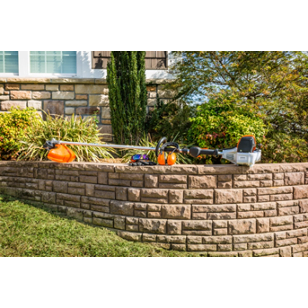 Professional straight shaft trimmer with orange and gray design resting on a stone retaining wall in a landscaped yard