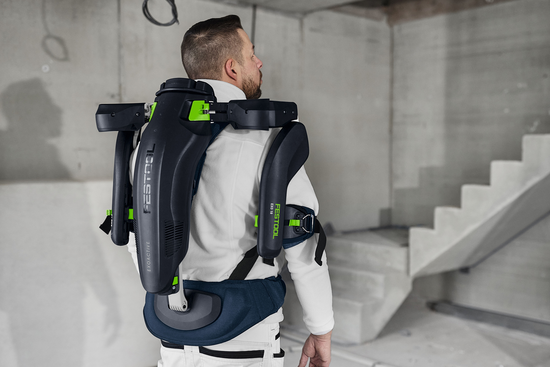 Man wearing black and green Festool ergonomic exoskeleton for upper back support in unfinished construction space
