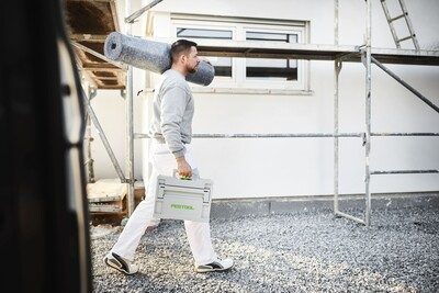 Man carrying compact Festool tool box and insulation roll at construction site with scaffolding and ladder in background