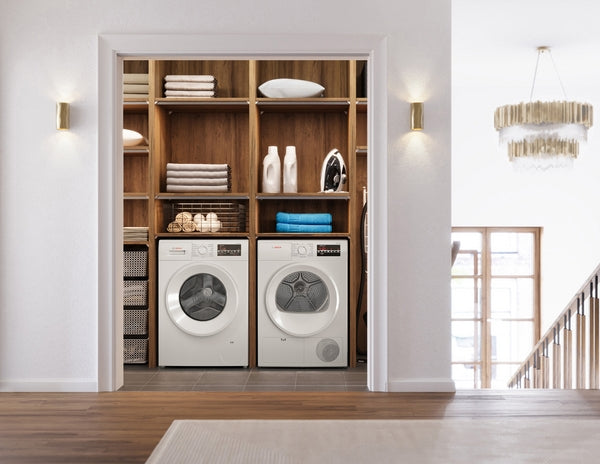 Laundry room with white front-loading washer and compact condensation dryer in wooden shelving unit under soft wall lights