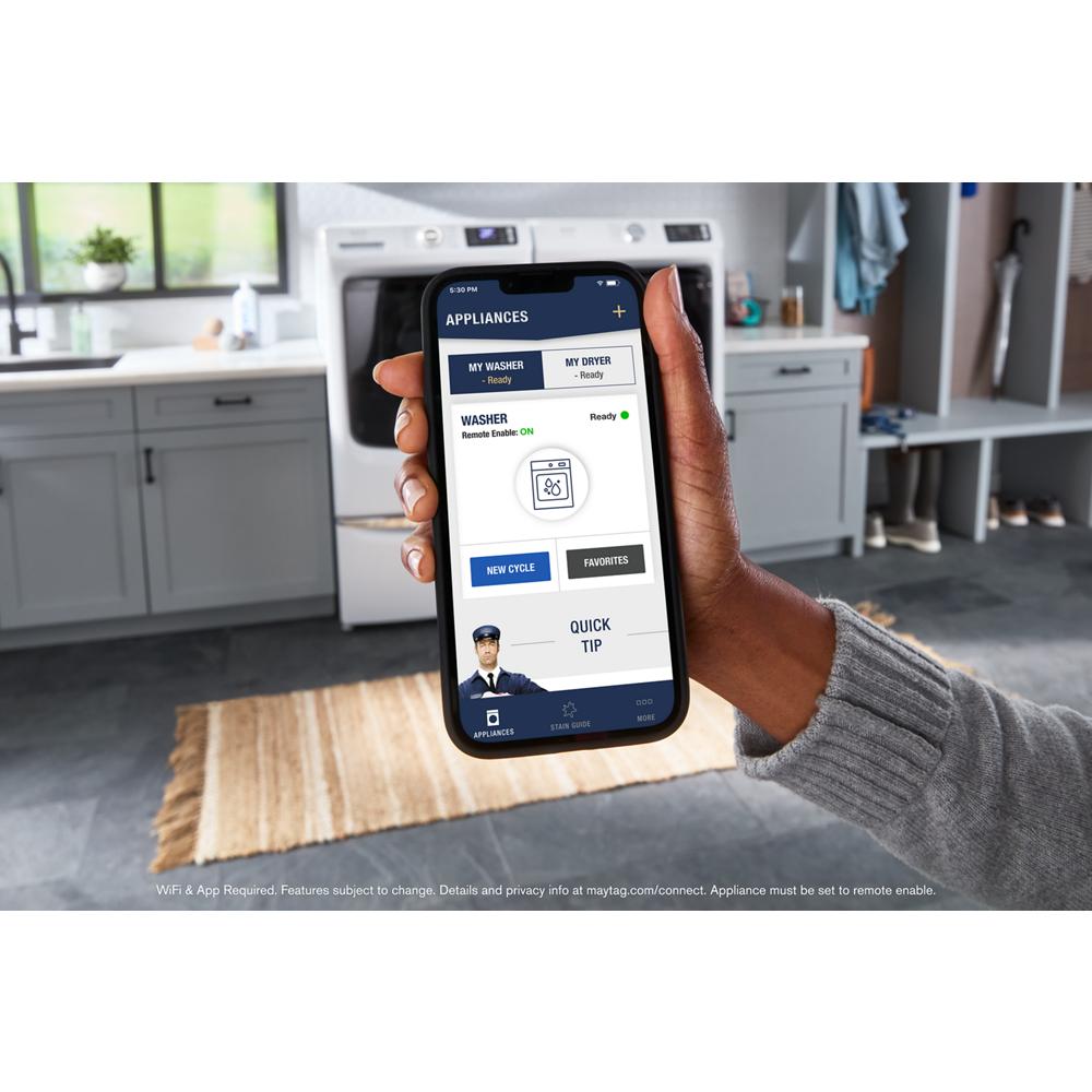 Hand holding smartphone with app controlling washer and dryer in modern laundry room with rug and bench background