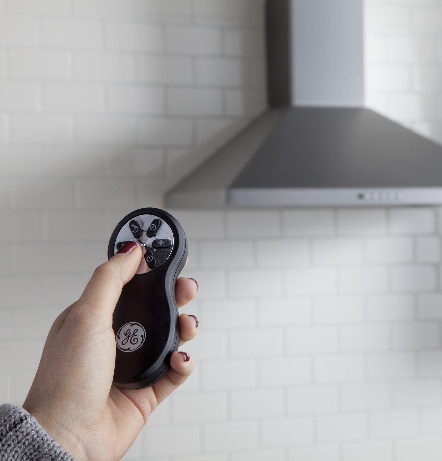 Hand holding remote control pointed at stainless steel wall-mounted pyramid chimney hood in kitchen setting