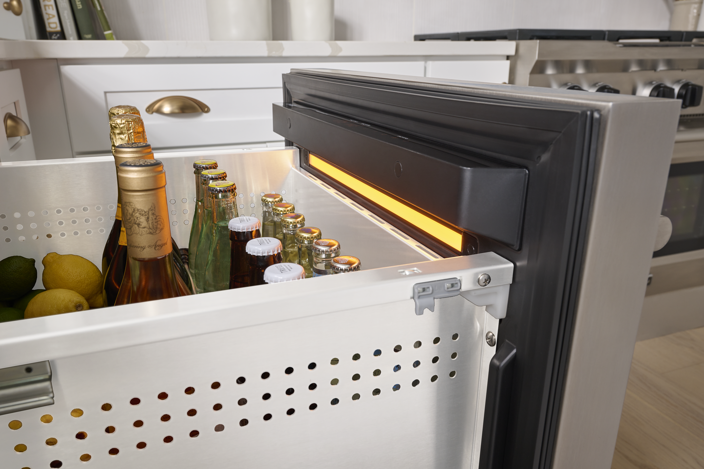 Under-counter stainless steel refrigerator drawer storing bottled beverages and citrus fruit in a modern kitchen environment