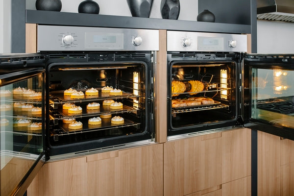 Two open stainless steel single wall ovens with right side doors baking cupcakes and rotisserie chicken in a modern kitchen