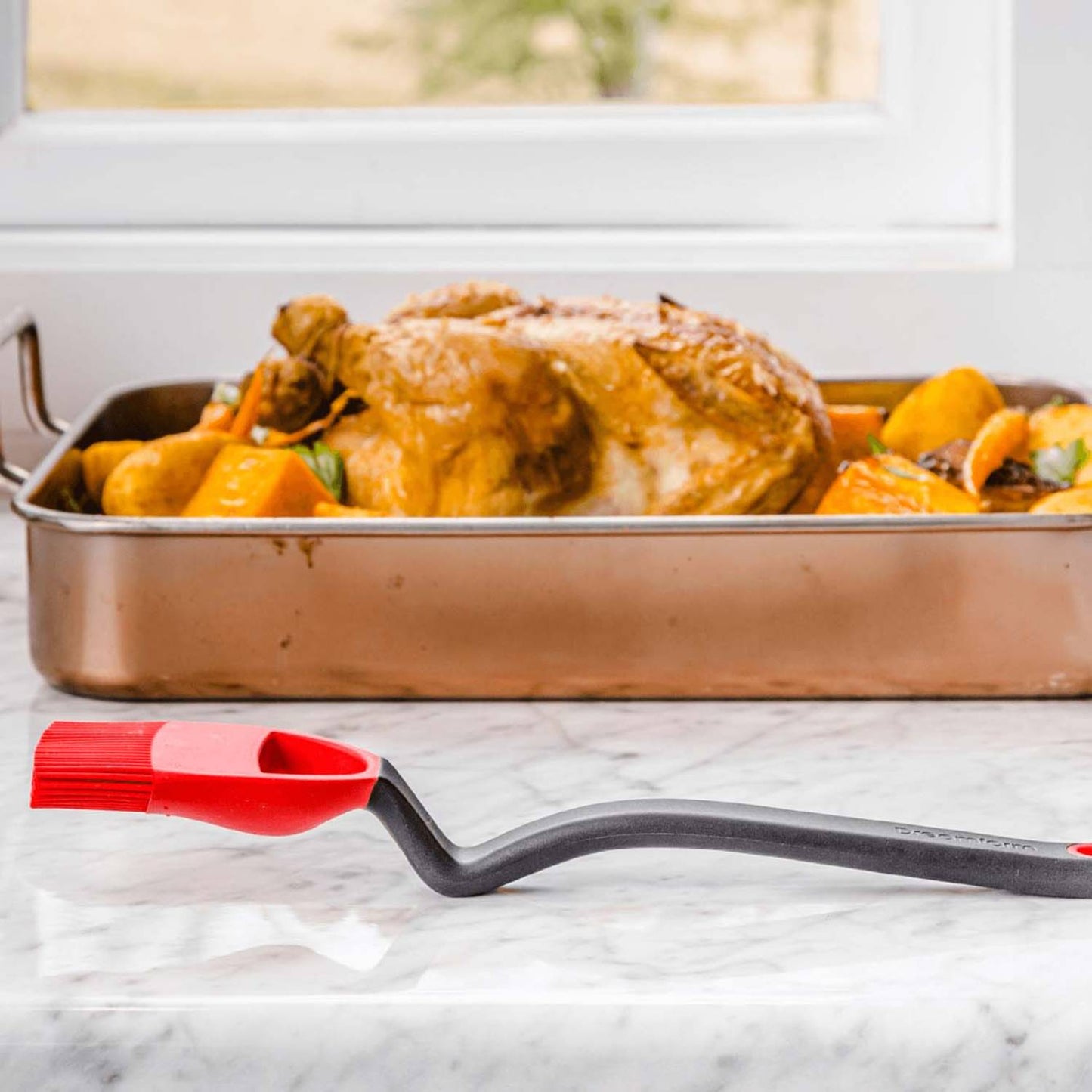 Red and black silicone basting brush with curved handle on marble surface next to roasted chicken in kitchenware collection