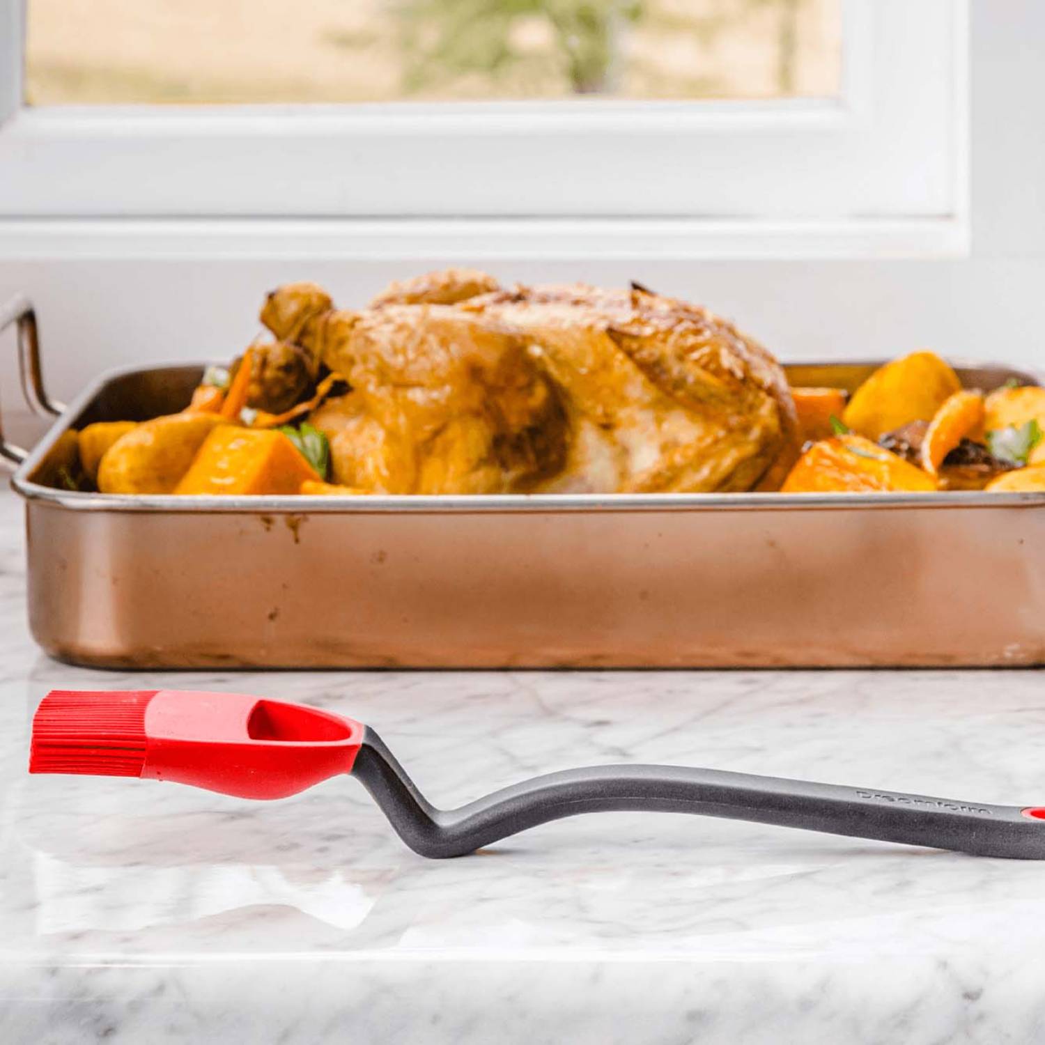 Red and black silicone basting brush with curved handle on marble surface next to roasted chicken in kitchenware collection