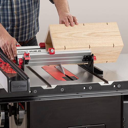 Hand operating Woodpeckers spline jig with red knobs guiding wooden box frame on table saw for joinery cuts