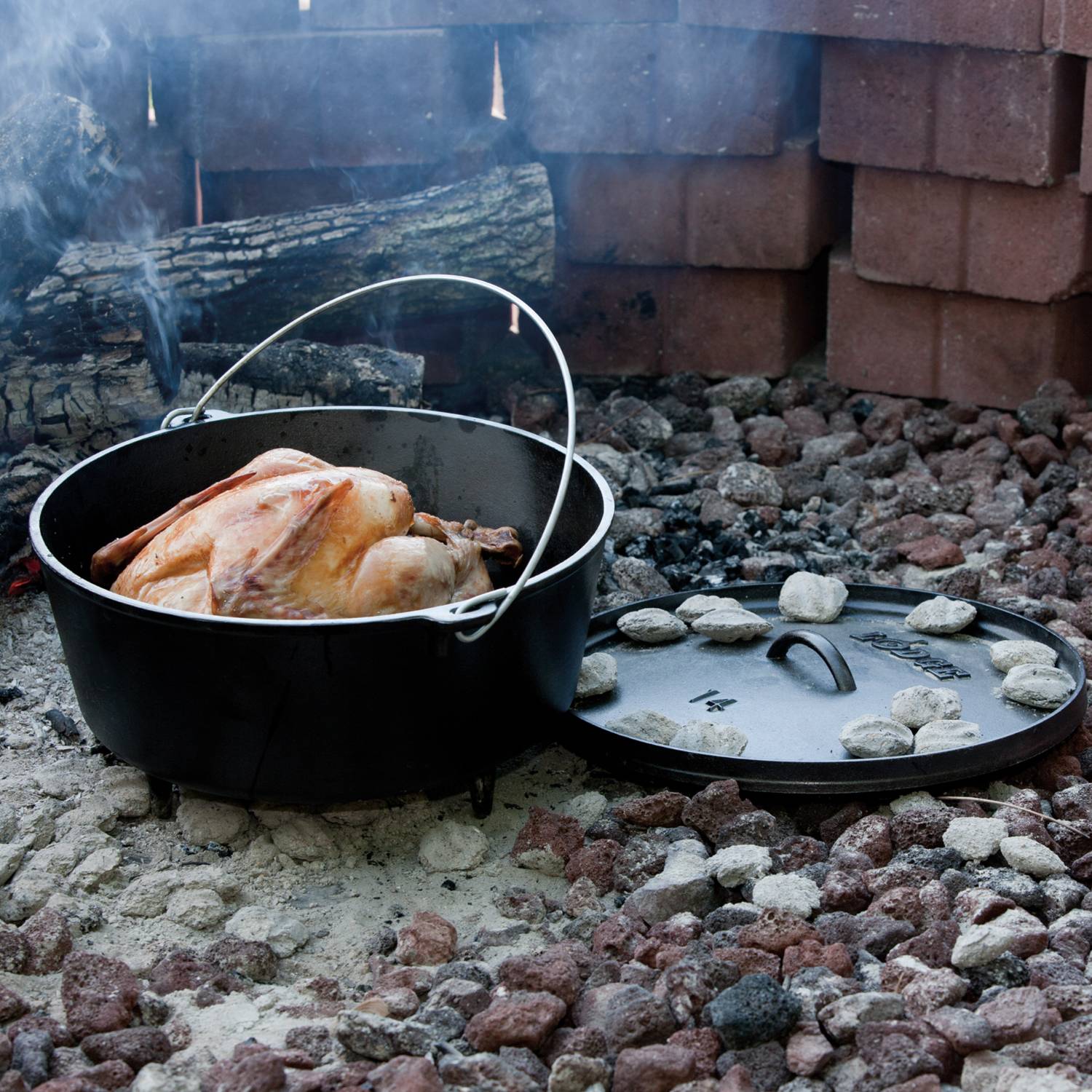 Cast iron camp Dutch oven cooking whole roasted chicken over hot coals with lid resting nearby in outdoor kitchenware setting