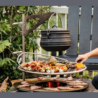 Cast iron kettle suspended over fire pit grill with skewers and food cooking in outdoor patio setting