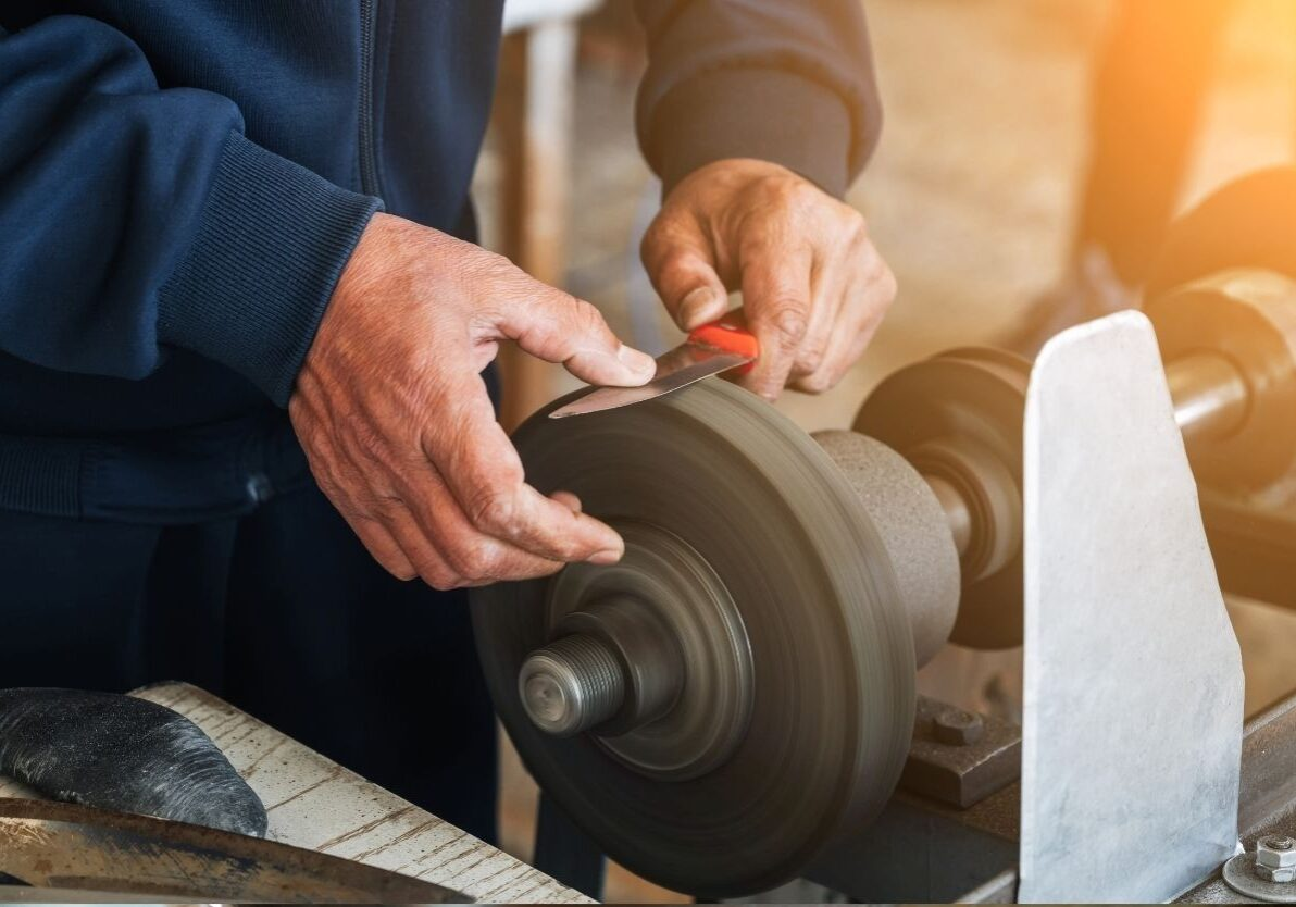 Close-up of a person sharpening a small knife on a rotating grinding wheel in a workshop bathed in warm natural light.