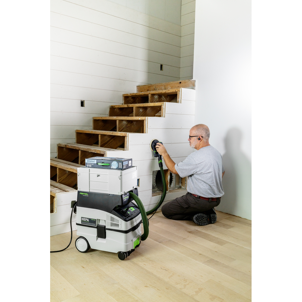 Man kneeling on hardwood floor sanding unfinished staircase with Festool power sander and dust extractor in home renovation setting
