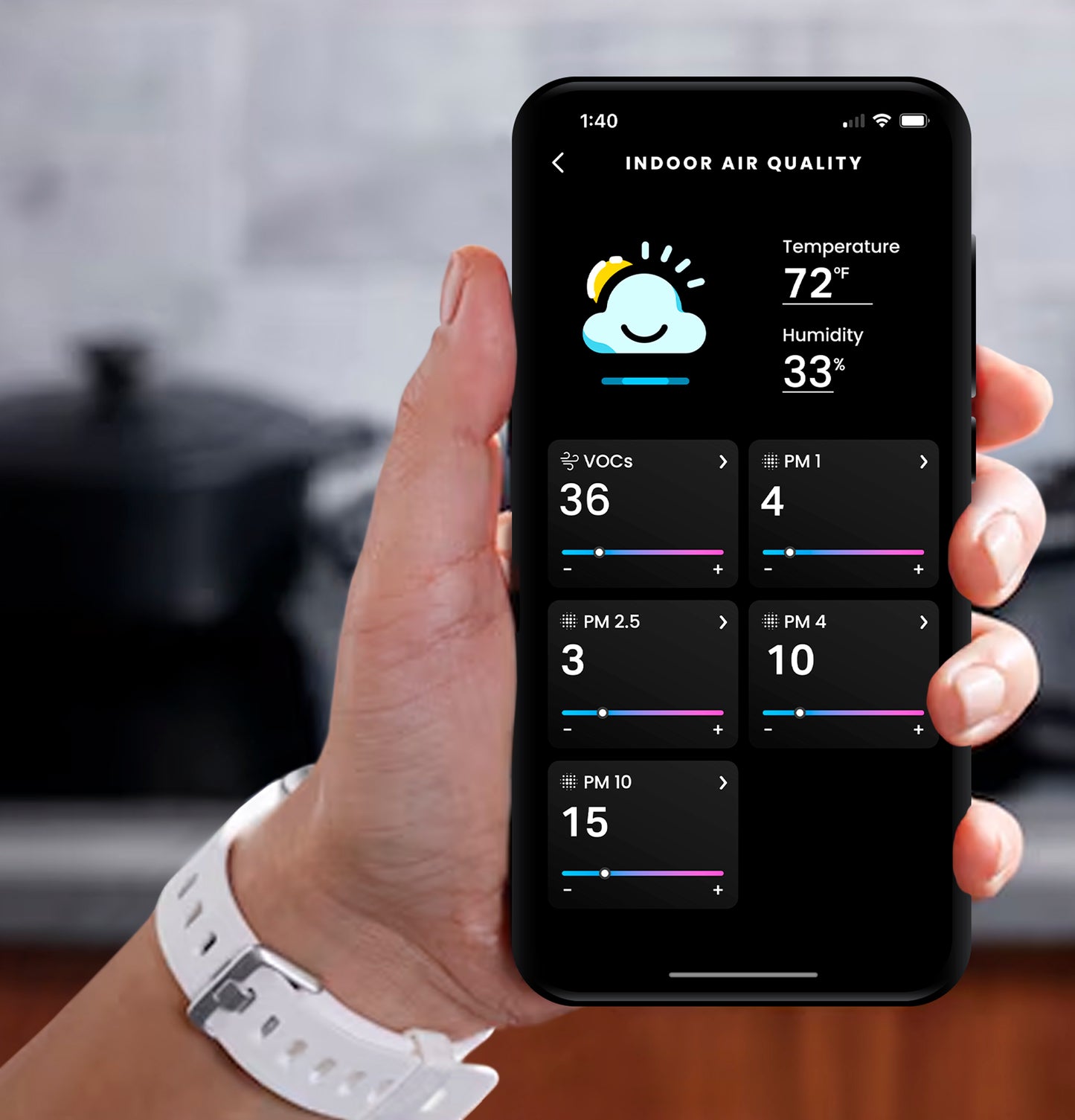 Hand holding smartphone displaying indoor air quality app with temperature, humidity, and pollutant levels in kitchen setting