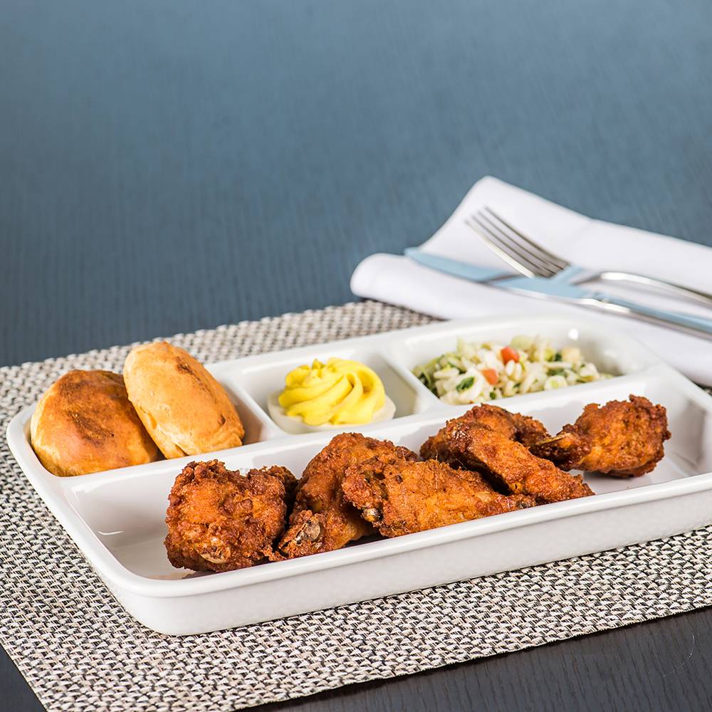 White TV dinner tray with four compartments holding fried chicken, dinner rolls, mashed potatoes, and salad on woven placemat