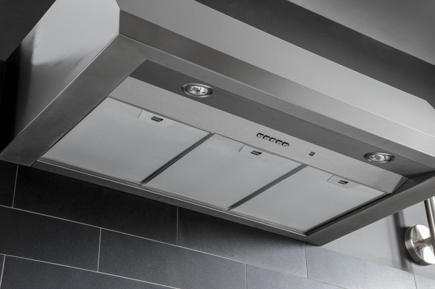 Stainless steel commercial style range hood with built-in lights mounted above dark tiled backsplash in kitchen appliances collection