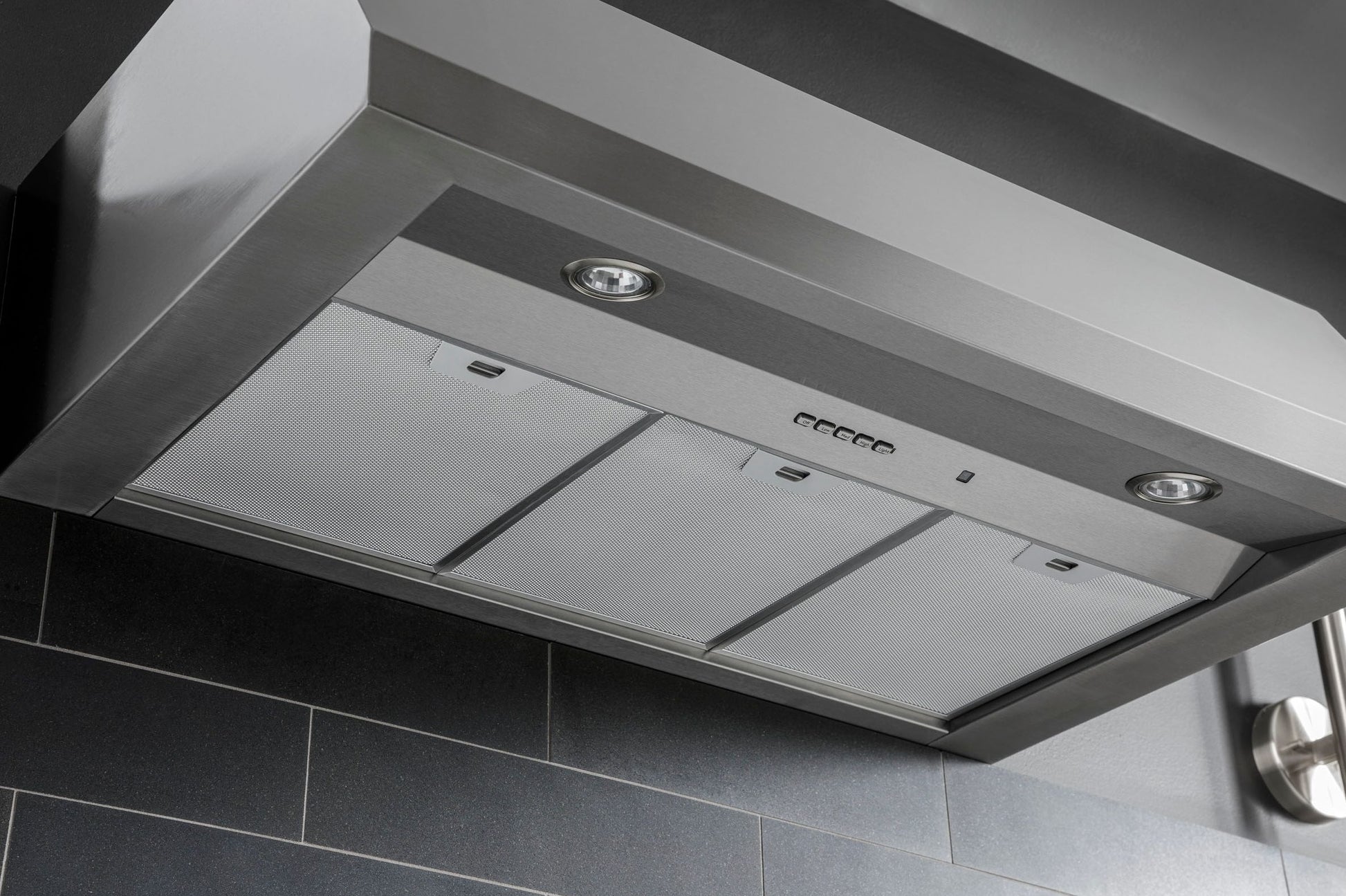 Stainless steel commercial style range hood with built-in lights mounted above dark tiled backsplash in kitchen appliances collection