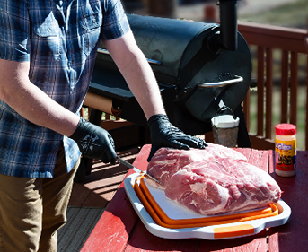 Person slicing a large cut of raw meat on an orange prep tub outdoors near a black grill for grilling tools use