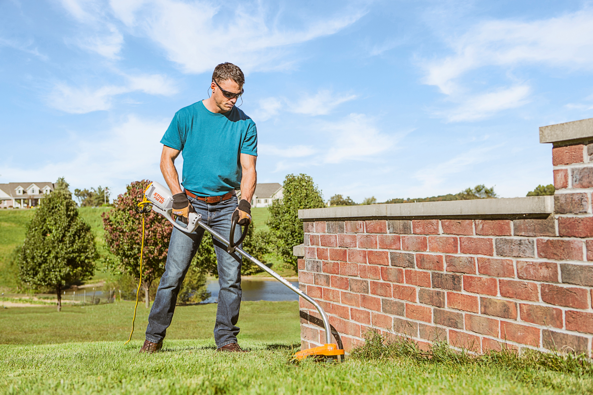 Man wearing protective glasses using STIHL electric trimmer to edge grass along a brick garden wall on a sunny day