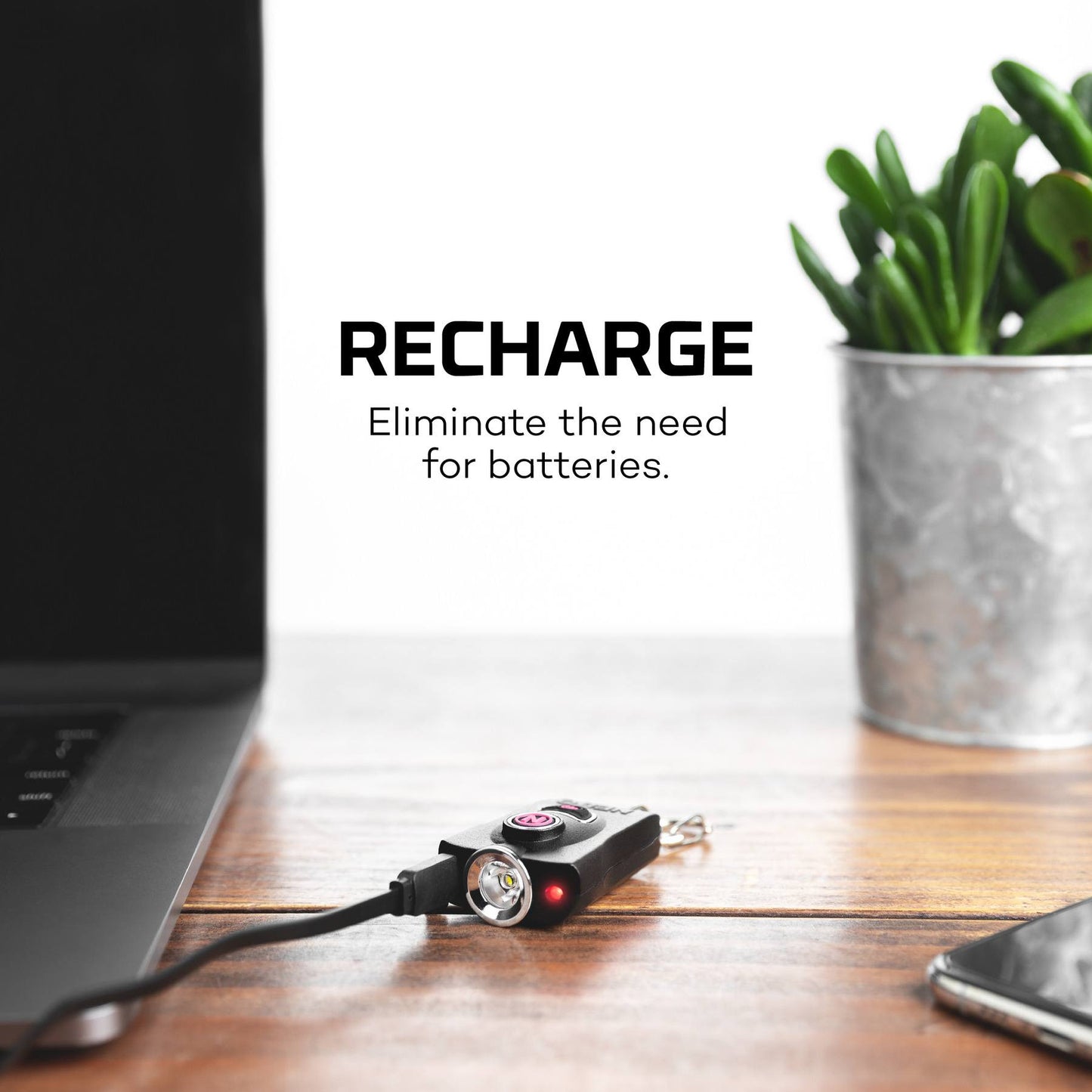 Black rechargeable keychain light and safety alarm plugged in on wooden desk with plant and laptop nearby