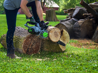 Person using green and gray cordless chainsaw to cut thick log outdoors for home improvement tools task