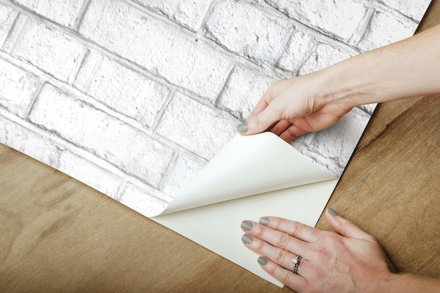 Hands applying whitewash brick pattern peel and stick wallpaper on wooden surface for wall decor.