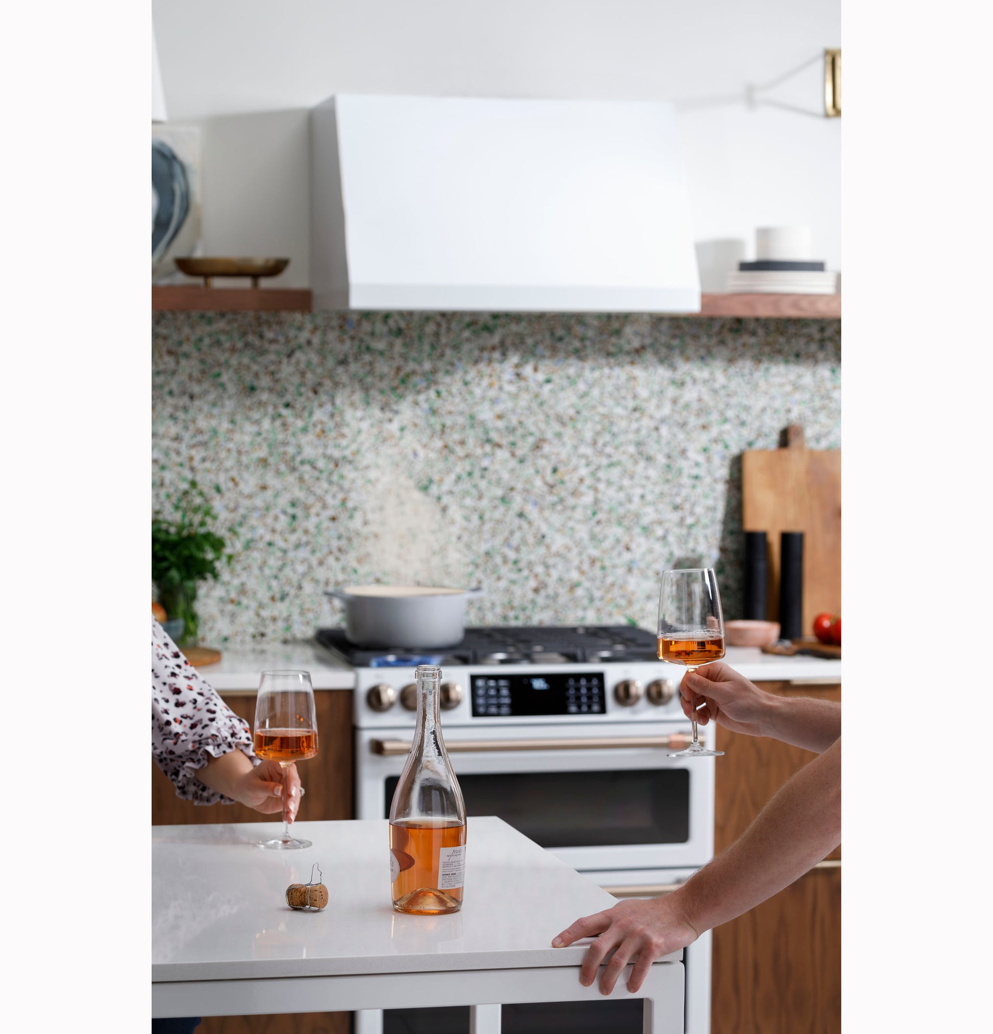 Modern kitchen scene with white commercial hood, terrazzo backsplash, stove, and two people holding wine glasses over island countertop