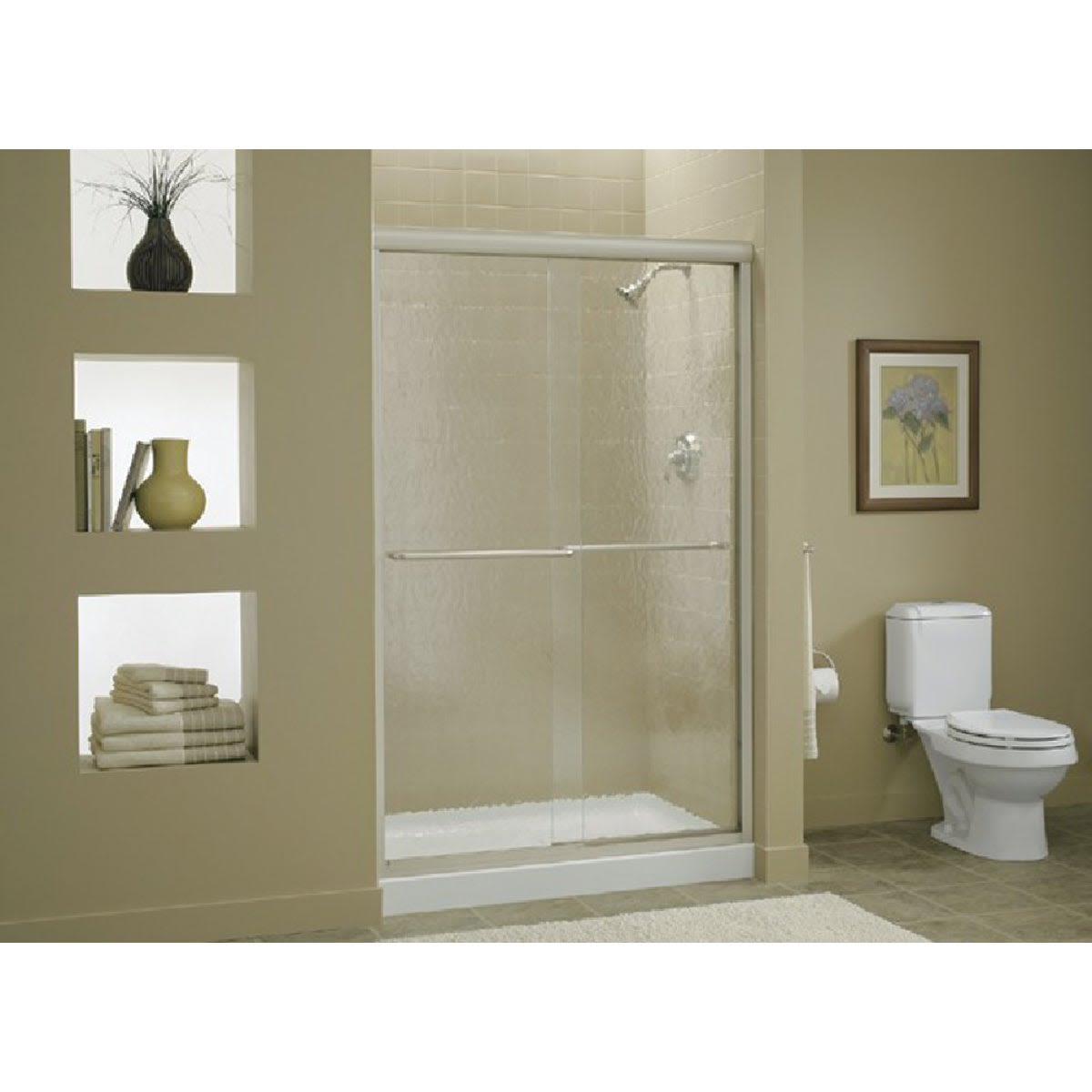 Clear sliding shower door with polished metal frame in a beige bathroom featuring decorative wall niches and folded towels