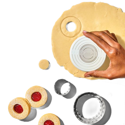 Hand holding round double-sided cookie cutter cutting dough with additional cutters and jam-filled cookies nearby