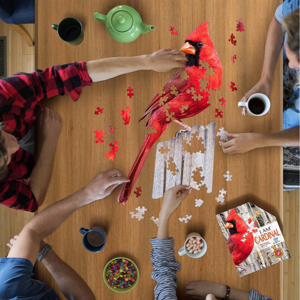People assembling a colorful cardinal-shaped 300-piece puzzle at a wooden table with drinks and snacks nearby
