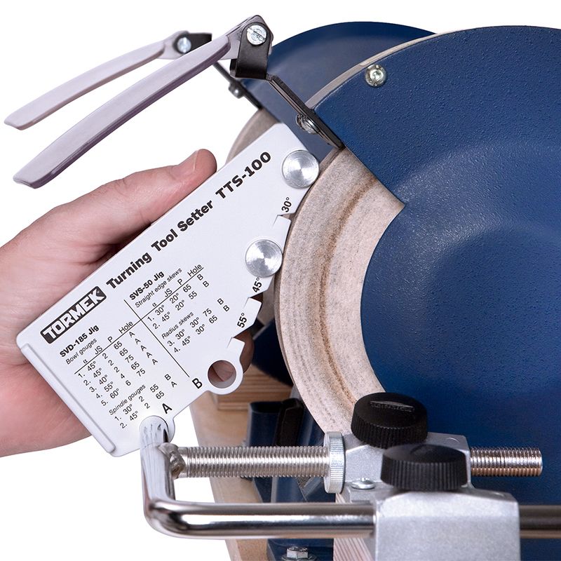 Hand holding Tormek TTS-100 tool setter against a blue grinding wheel for precise sharpening adjustments
