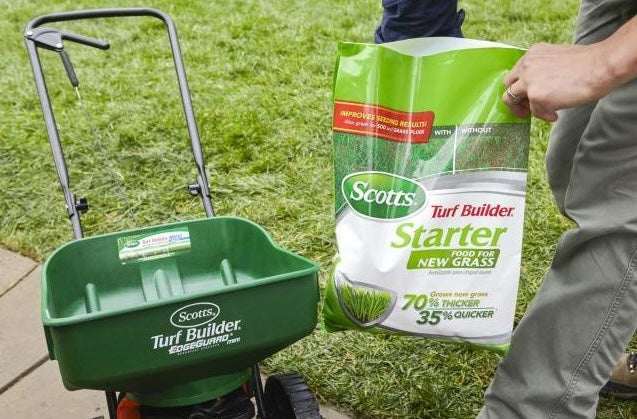 Bag of starter fertilizer being poured into a green Scotts Turf Builder spreader on a grass lawn