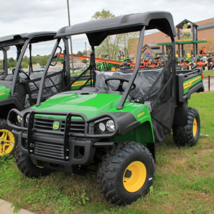 Green utility vehicle with protective roof and off-road tires parked on grass near outdoor equipment display