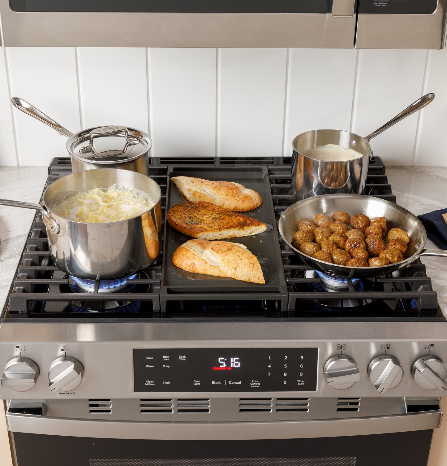 Modern stainless steel gas range with pots cooking pasta, sauce, meatballs, and bread on griddle in kitchen setting