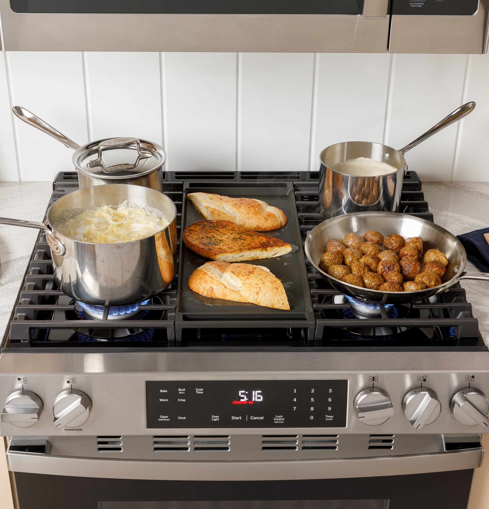Modern stainless steel gas range with pots cooking pasta, sauce, meatballs, and bread on griddle in kitchen setting