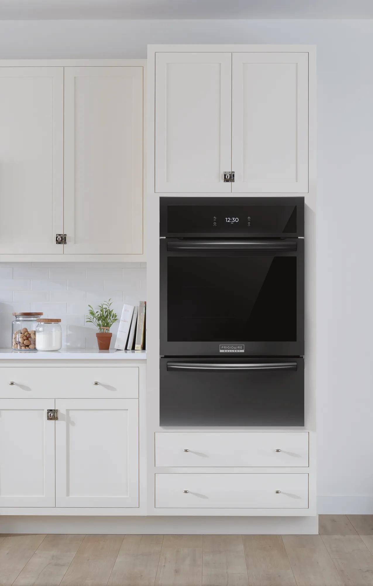 Built-in black gas wall oven with digital clock and handle, integrated into white kitchen cabinetry with drawers and cupboards.
