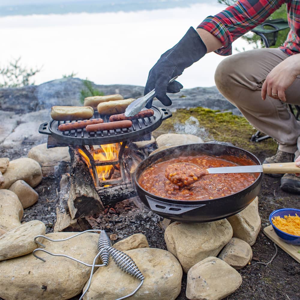 Outdoor campfire cooking with cast iron grill toasting buns and skillet simmering chili on rocks in nature setting
