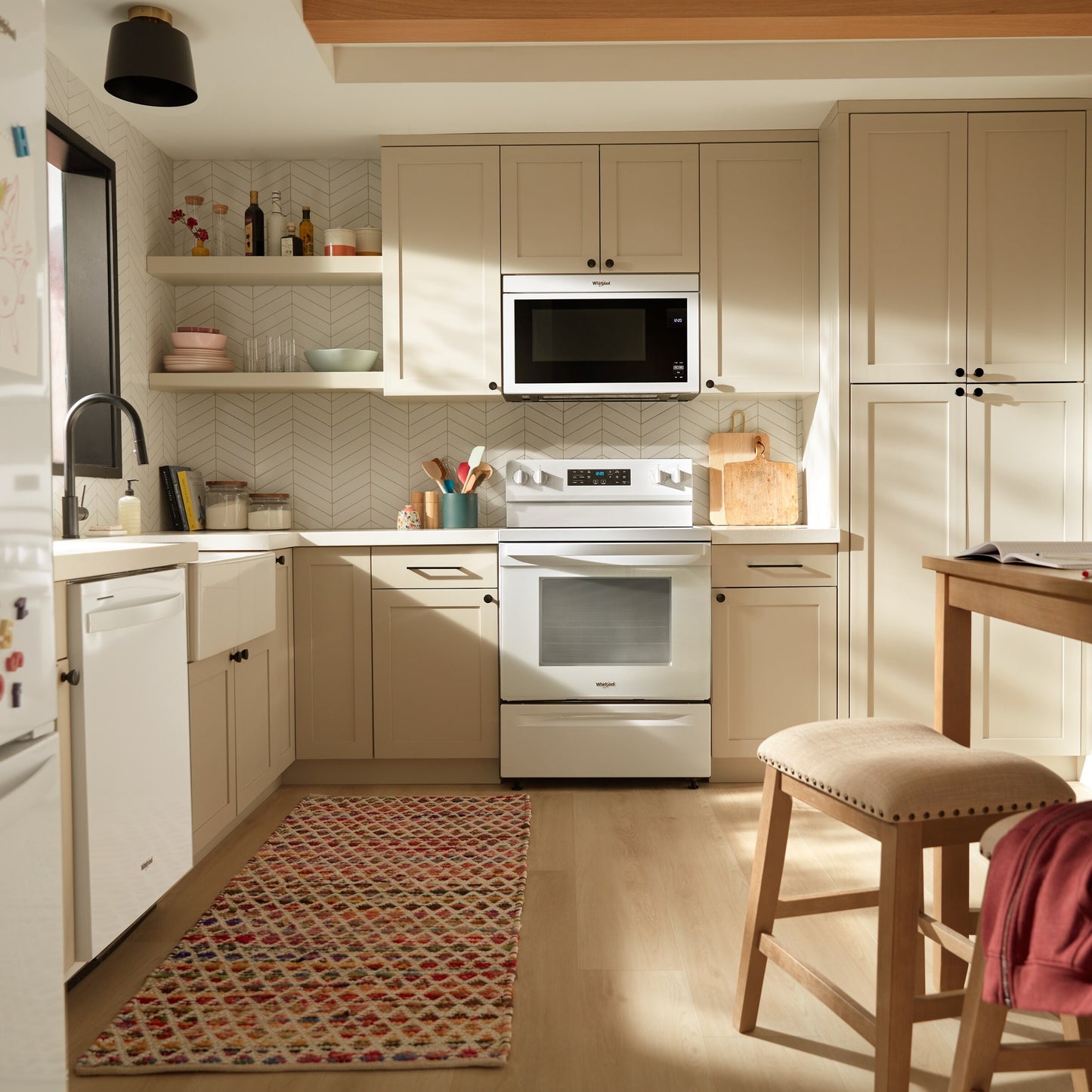 Bright kitchen with white electric range, microwave, farmhouse sink, beige cabinets, and patterned rug on light wood floor