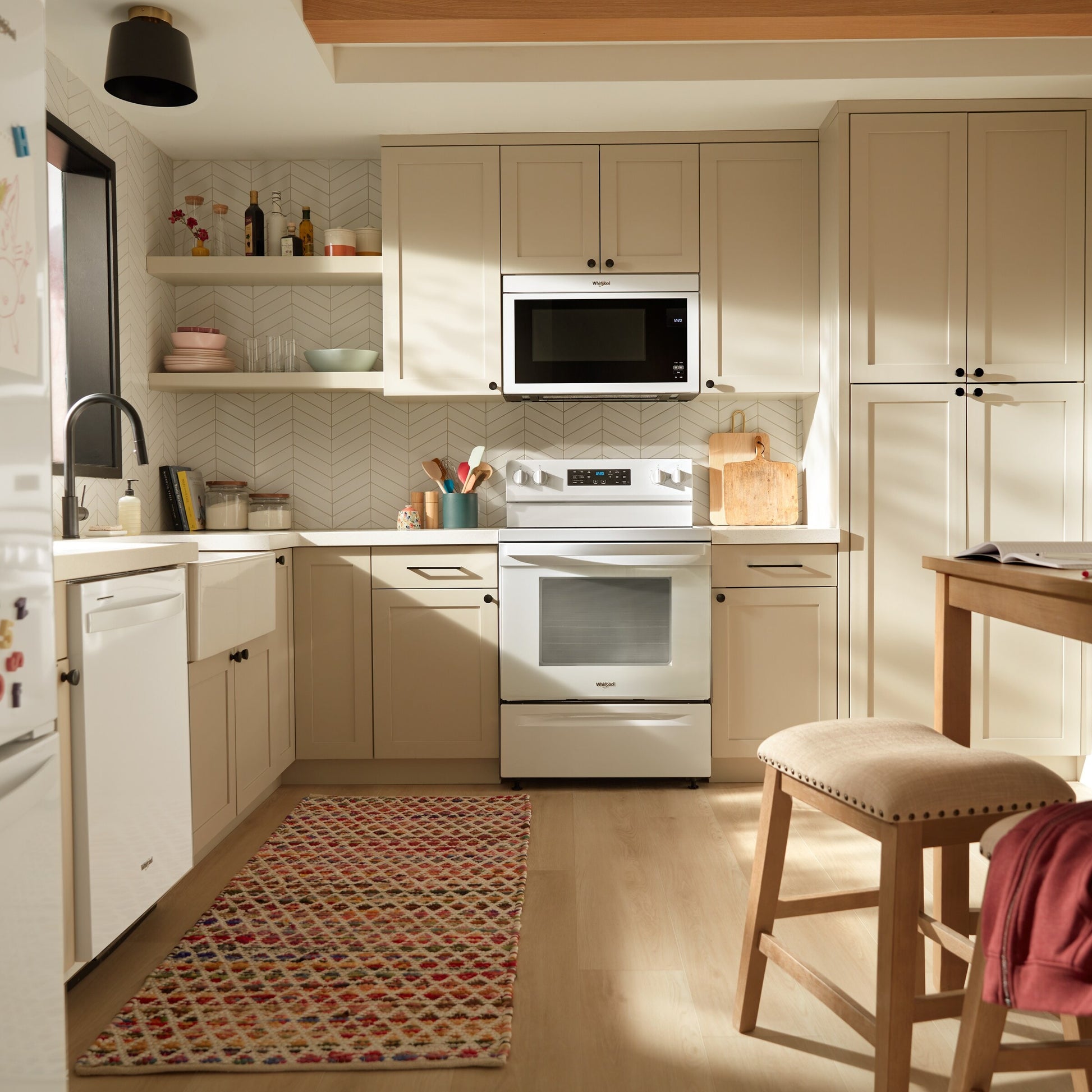 Bright kitchen with white electric range, microwave, farmhouse sink, beige cabinets, and patterned rug on light wood floor