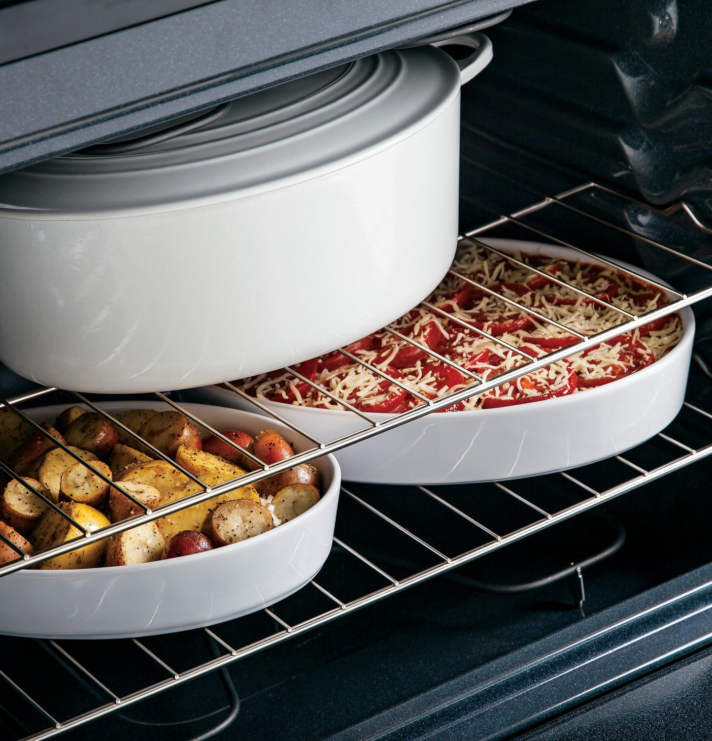 White ceramic dishes with potatoes, pizza, and a covered pot inside a built-in oven rack in kitchen appliance setting