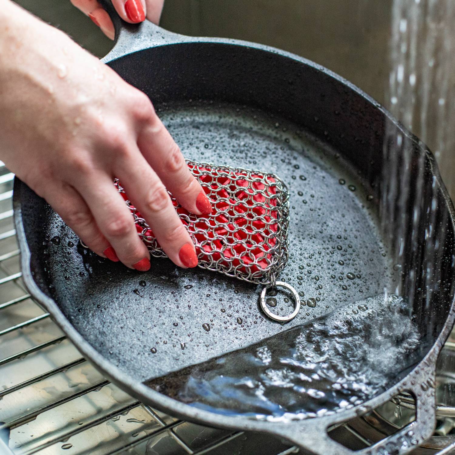Hand using red chainmail scrubbing pad to clean cast iron skillet under running water in kitchenware setting