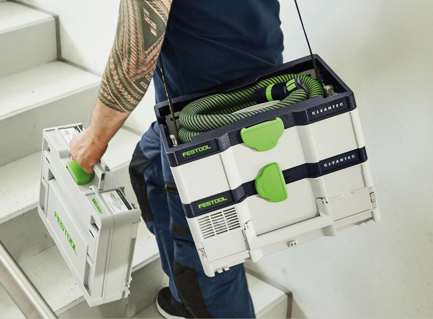 Person carrying green and white cordless mobile dust extractor and a matching Festool tool case up indoor stairs