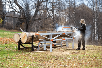 Outdoor woodworking setup featuring a portable saw table with orange clamps cutting large logs in a wooded area
