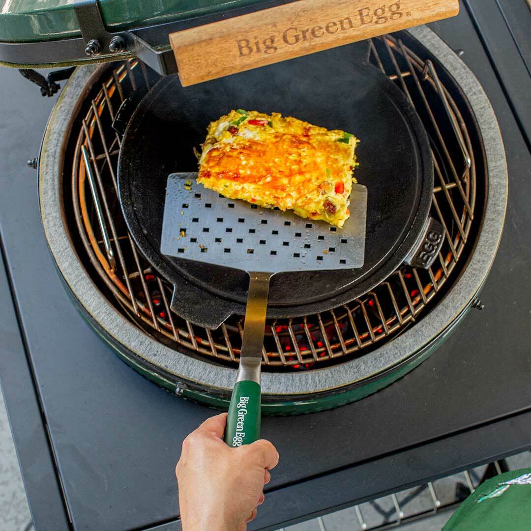 Hand holding stainless steel wide spatula with green handle lifting grilled vegetable frittata on outdoor ceramic grill