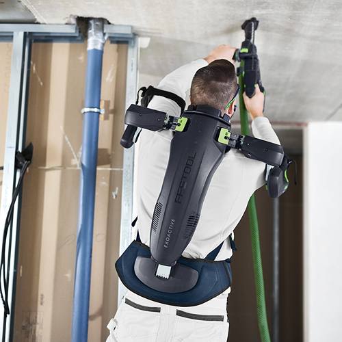 Person wearing black and green ergonomic exoskeleton supporting upper back and shoulders while using power tool indoors
