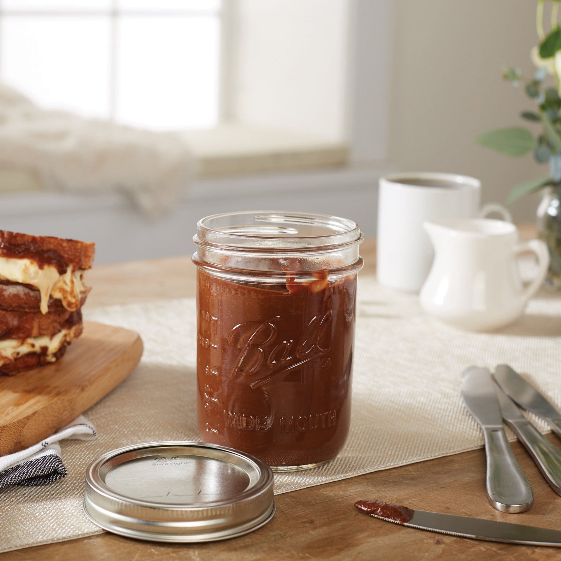 Clear glass wide mouth mason jar filled with chocolate spread beside grilled cheese sandwich and kitchen utensils on table