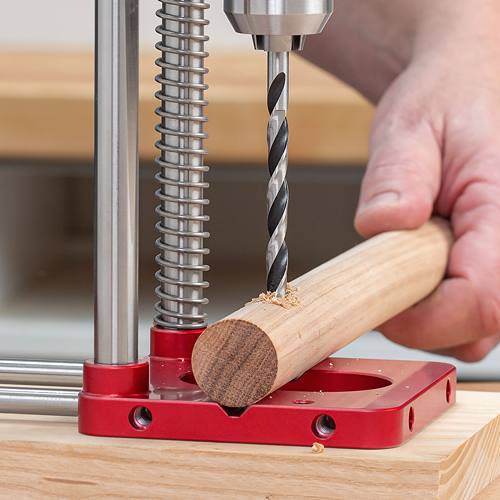 Red metal drill guide with spring mechanism securing round wooden dowel for precise holes in tool accessories collection